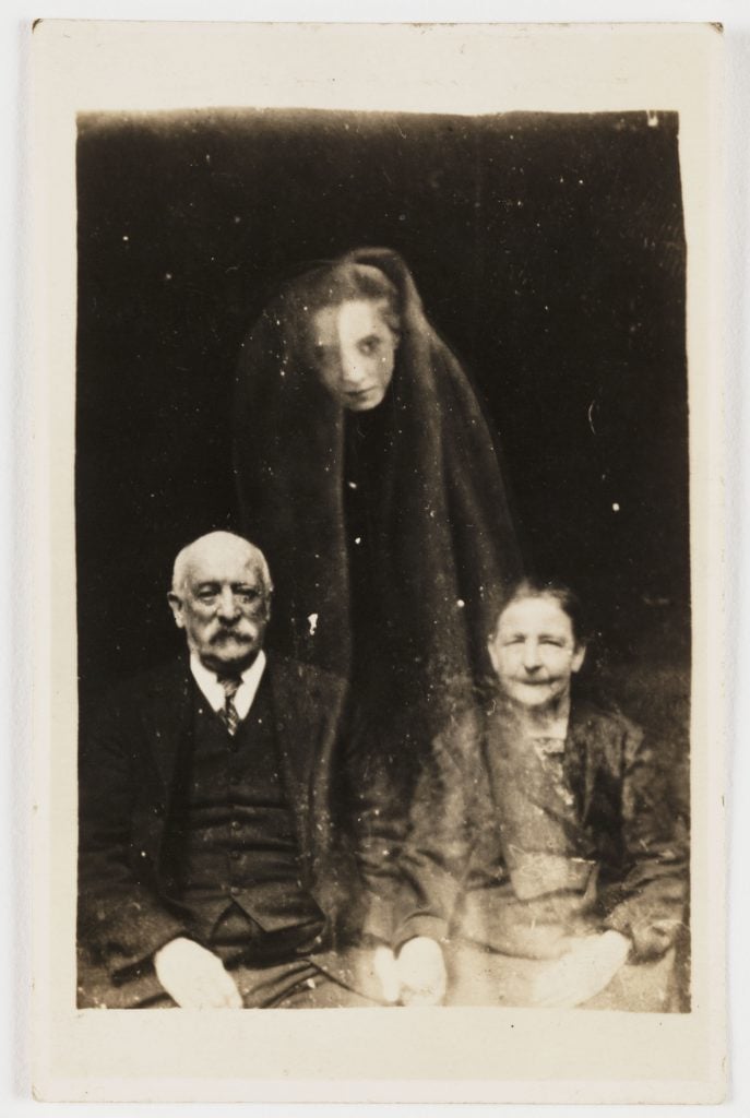 The face of a young woman appears as if floating above a seated elderly couple, wrapped in a cloak.